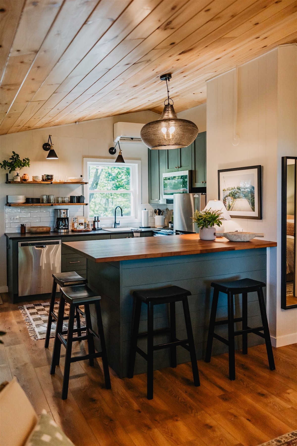 The kitchen features polished wooden surfaces and modern amenities. A central bar-height island offers seating with three black stools. Natural light enters through a nearby window, highlighting the tasteful decor and thoughtfully organized shelves filled with kitchen essentials.
