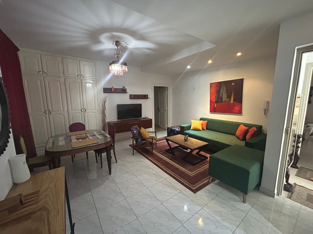 A living area features a green sectional sofa with colorful cushions and a wooden coffee table. A dining table with chairs is positioned nearby. A television is mounted on a light-colored cabinet, and a piece of artwork hangs on the wall, highlighting the space's clean design.