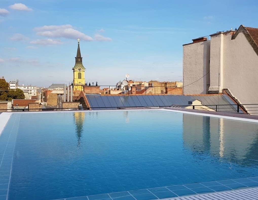 A rooftop pool offers a clear view of the skyline, with a clock tower visible in the distance. The water's surface reflects the sky, and the surrounding area features various rooftops, showcasing the blend of urban architecture.