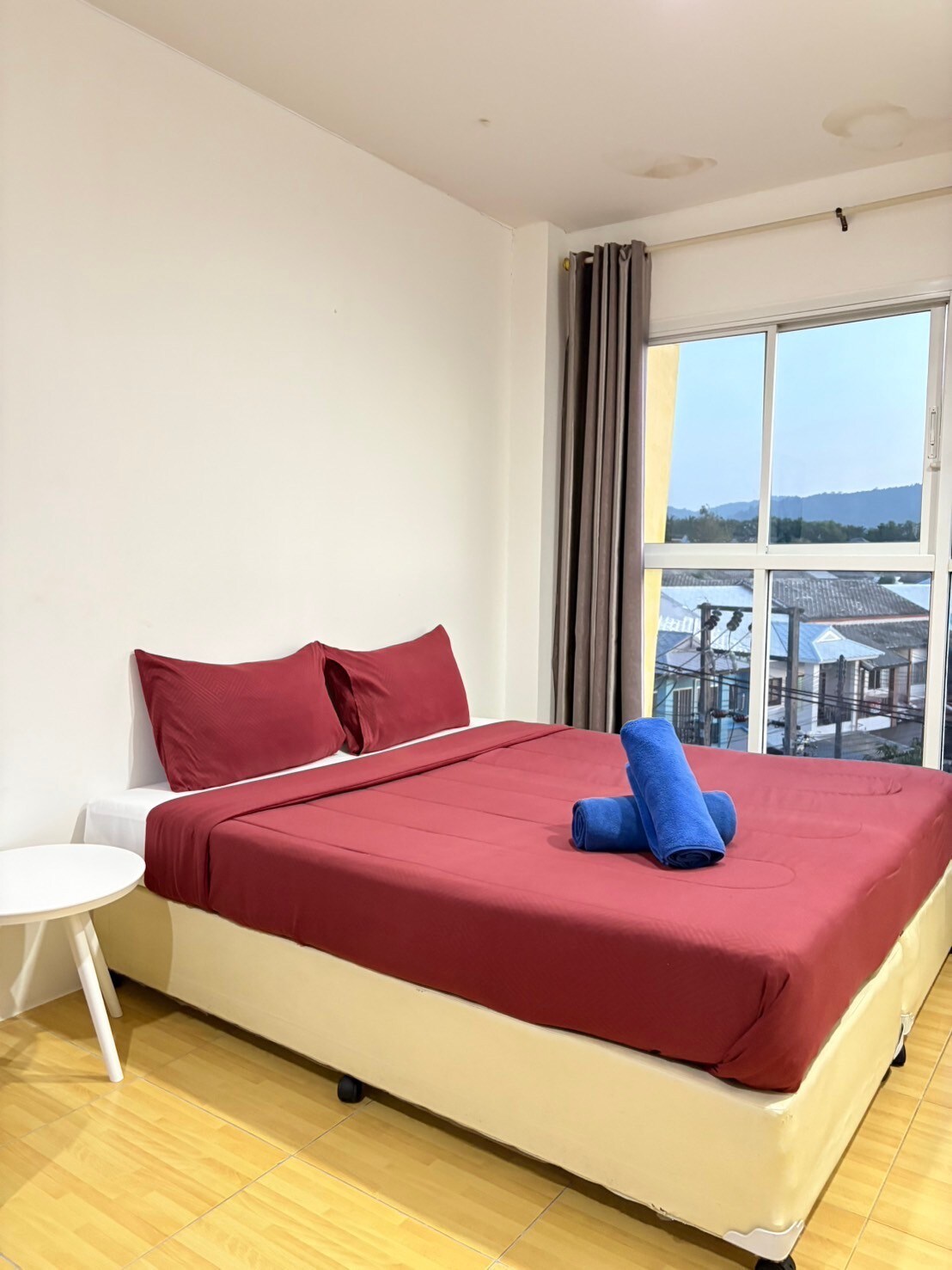 A modern room is highlighted by a queen-sized bed with burgundy linens and matching towels. A small white side table is positioned beside the bed. Soft light enters through a large window framed with light curtains, offering views of the surroundings.