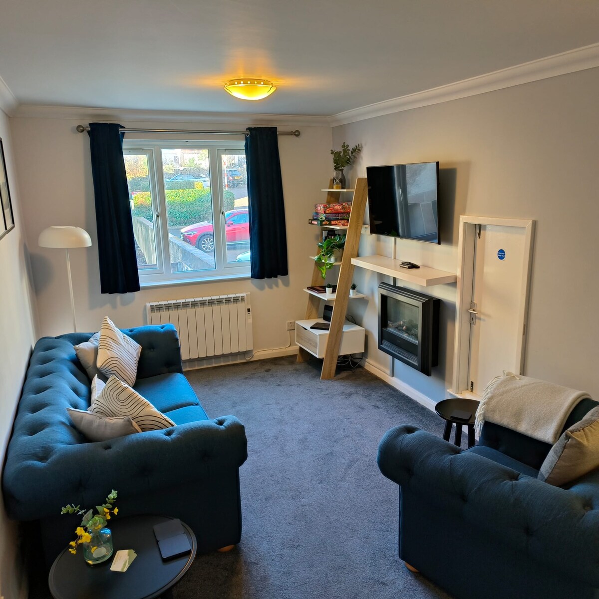 A cozy lounge area features two blue couches arranged around a small table. A flat-screen TV is mounted on the wall across from the seating. A window with blue curtains allows natural light to fill the space, enhancing the relaxed atmosphere.