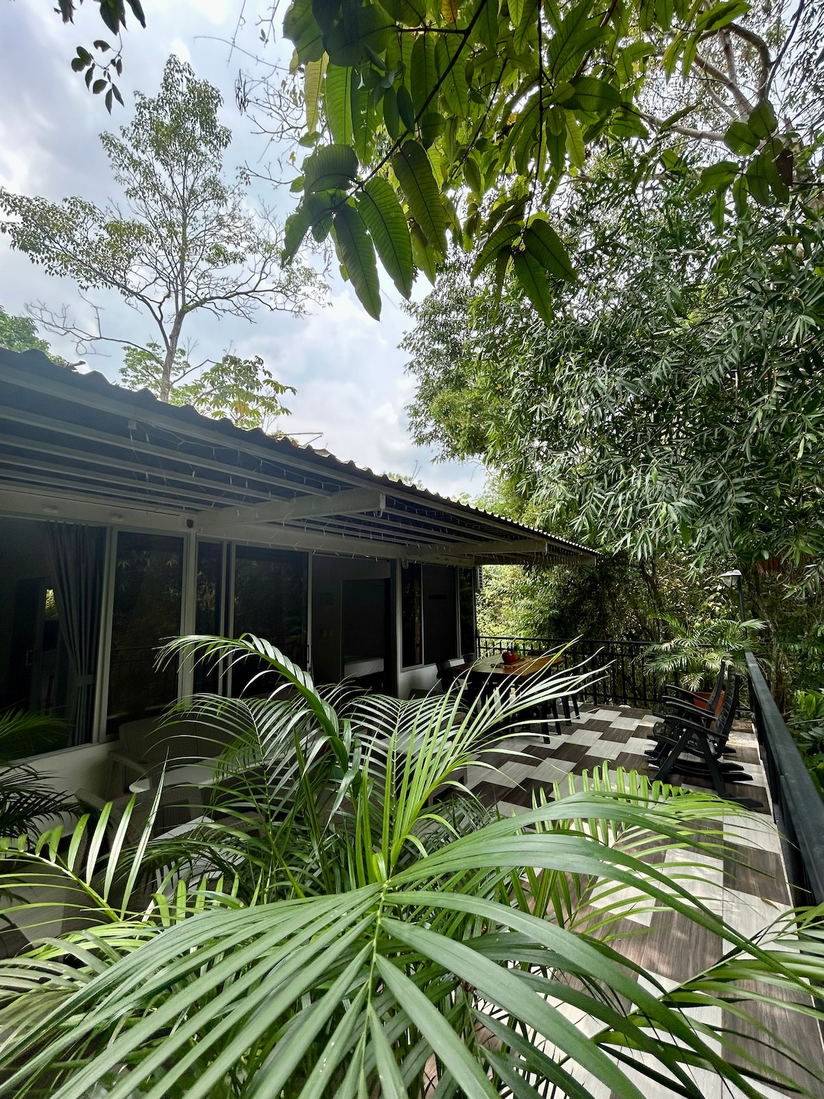 The exterior of the cabin is surrounded by lush green foliage, featuring a spacious patio with wooden furniture. Large windows reflect the natural light, while a corrugated roof provides shade. The tranquil setting showcases a blend of nature and modern design.