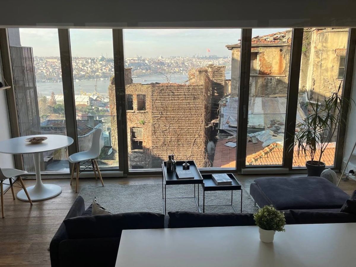 A modern living area is highlighted by large glass windows offering a sweeping view of the Golden Horn and old Istanbul. A comfortable sofa faces the vista, while a round dining table sits nearby. Natural light floods the space, enhancing the minimalist decor.