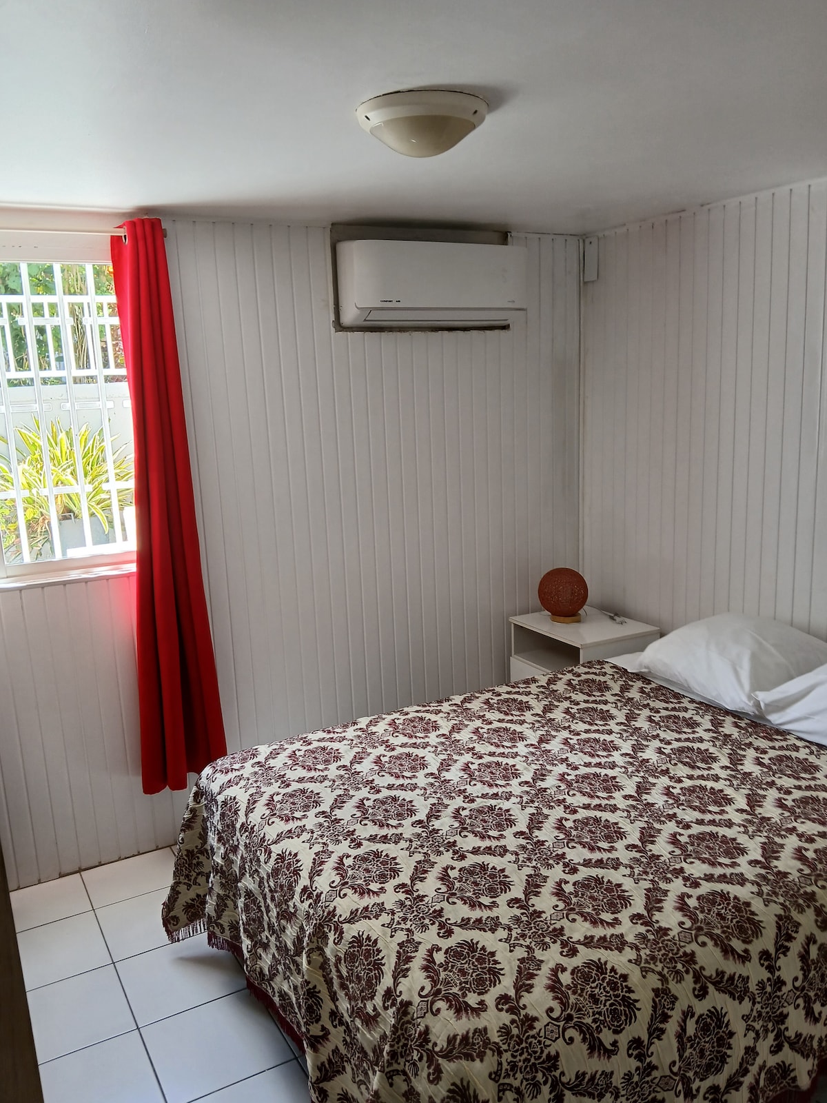 A cozy bedroom features a bed with a patterned coverlet, complemented by a bedside table holding a decorative item. Air conditioning is provided for comfort. Natural light filters through a window adorned with red curtains, creating a calm and inviting environment.