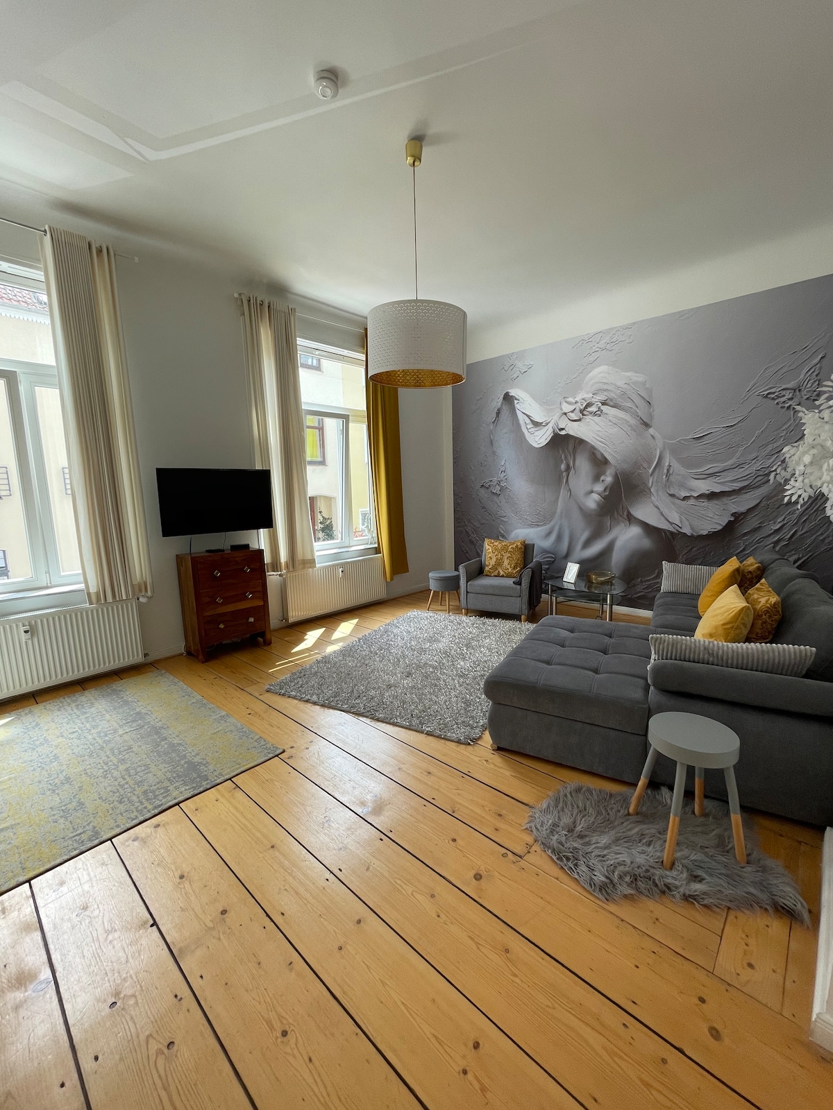 The bright living area features large windows allowing natural light to fill the space. A grey sectional sofa is paired with a small side table and decorative pillows. A modern light fixture hangs above, and a striking wall mural adds character, complemented by a wooden floor.