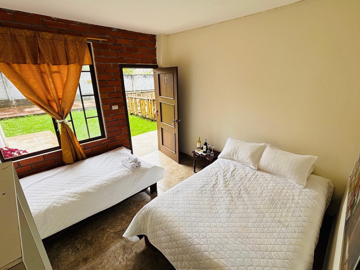 A cozy bedroom features a spacious bed alongside a twin bed, with natural light streaming through a large window. A wooden door opens to a lush green outdoor area, complementing the warm tones of the brick wall and soft bedding.