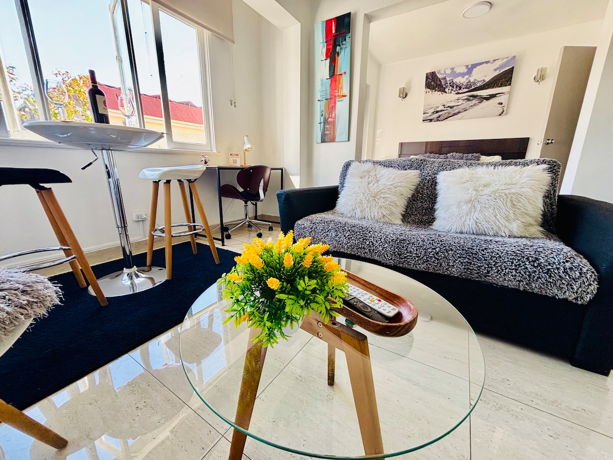 A living area is depicted with a modern black sofa featuring cozy throw pillows. A glass coffee table with decorative flowers sits in front. A dining table and chair are visible by the window, allowing natural light to fill the space. Artwork hangs on the wall, enhancing the ambiance.