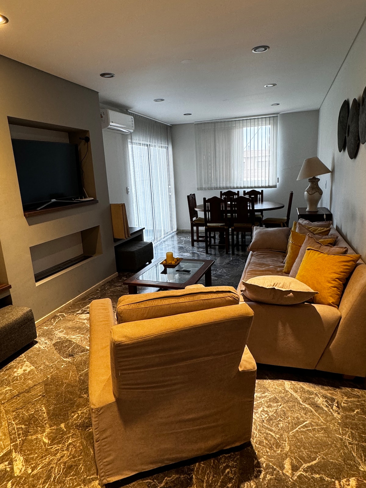 A spacious living area features a comfortable seating arrangement with light brown sofas and decorative cushions. A wooden dining table with chairs is positioned nearby. Natural light filters in through sheer curtains, and a television is mounted on the wall, creating a relaxed atmosphere.