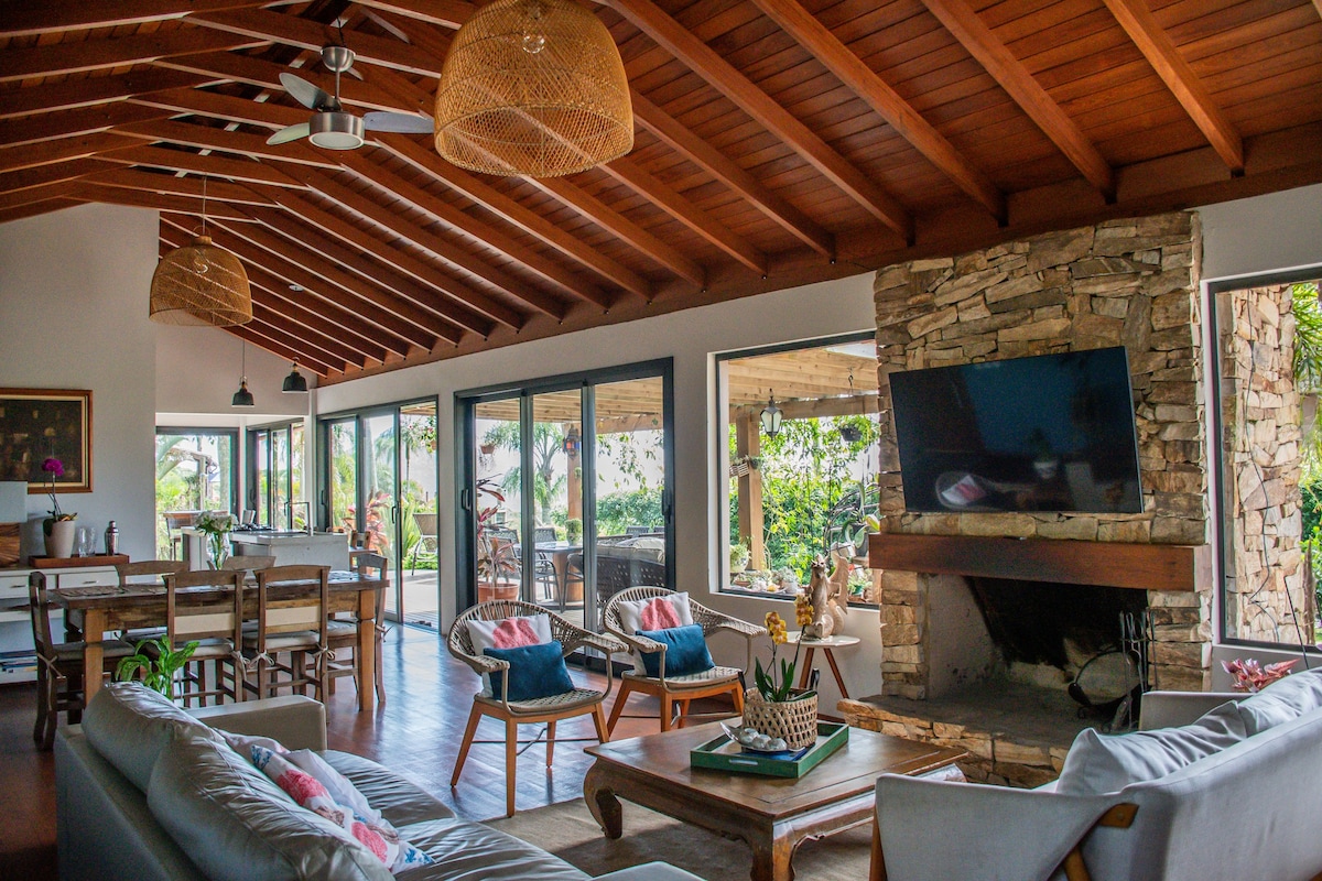 An open living area features vaulted wooden ceilings and large glass doors, allowing natural light to fill the space. A stone fireplace serves as a focal point, while comfortable seating arrangements are positioned around a wooden coffee table. Dining and outdoor seating areas are visible in the background.