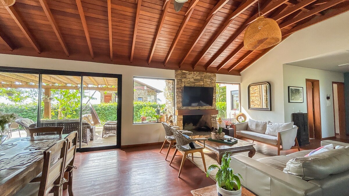 A living and dining area features wood-beamed ceilings and large glass doors, providing a view of the outdoor space. A stone fireplace serves as a focal point, with comfortable seating and a dining table nearby. Natural light fills the room, enhanced by plants and decorative elements.