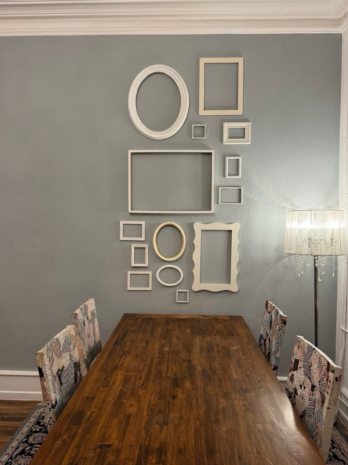 A long wooden dining table is positioned in front of a wall adorned with various empty picture frames in different shapes and sizes. A soft, warm light is emitted from a nearby floor lamp, creating a cozy ambiance in the space.
