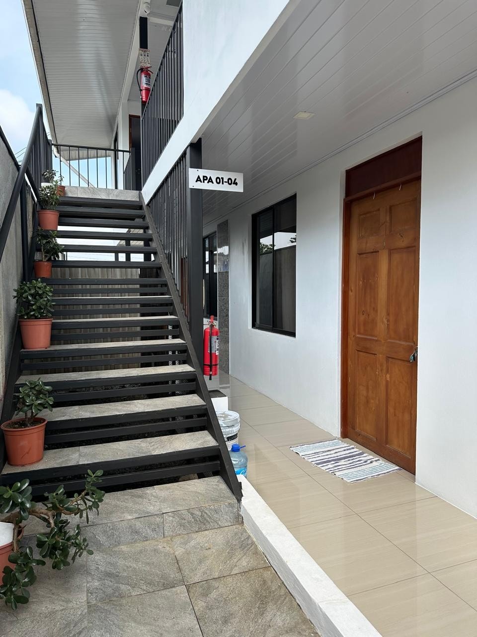 The exterior features a stairway with black metal railings leading to a second level. Potted plants are placed along the base, and a wooden door marked 'APA 01-04' is visible. The tiled floor reflects natural light, providing a clean and welcoming entryway.