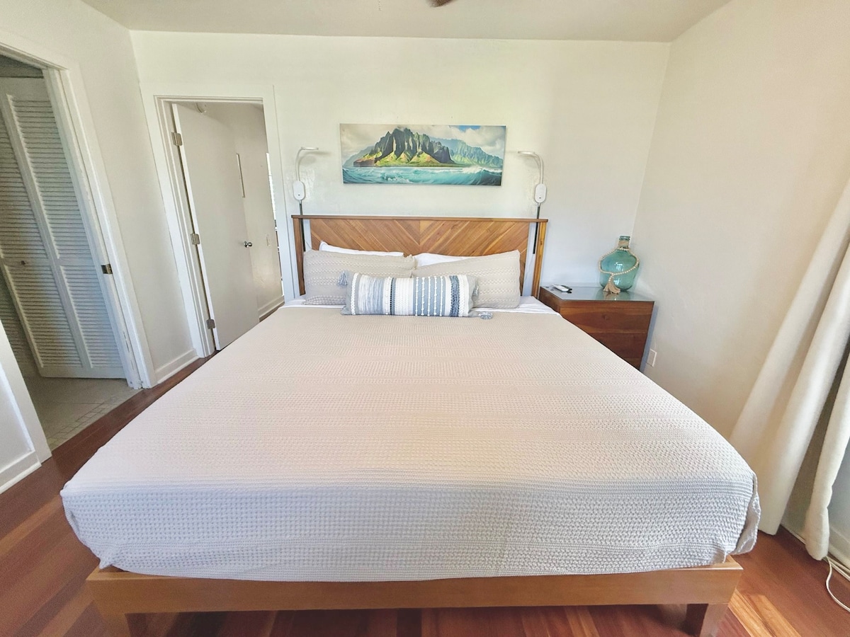 A spacious sleeping area features a king-size bed with a textured white duvet. On the wall above, a colorful landscape painting is displayed. A nightstand with a decorative lamp is positioned alongside the bed, and natural light filters through the curtains.