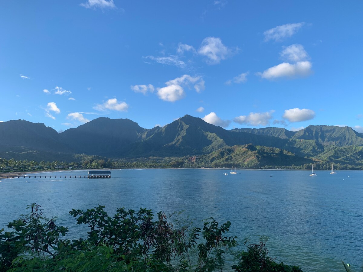 A scenic view of lush mountains rising dramatically above a calm ocean. Sailboats are gently anchored in the water, and a pier extends into the bay. The sky is clear with a few scattered clouds, enhancing the natural beauty of the landscape.