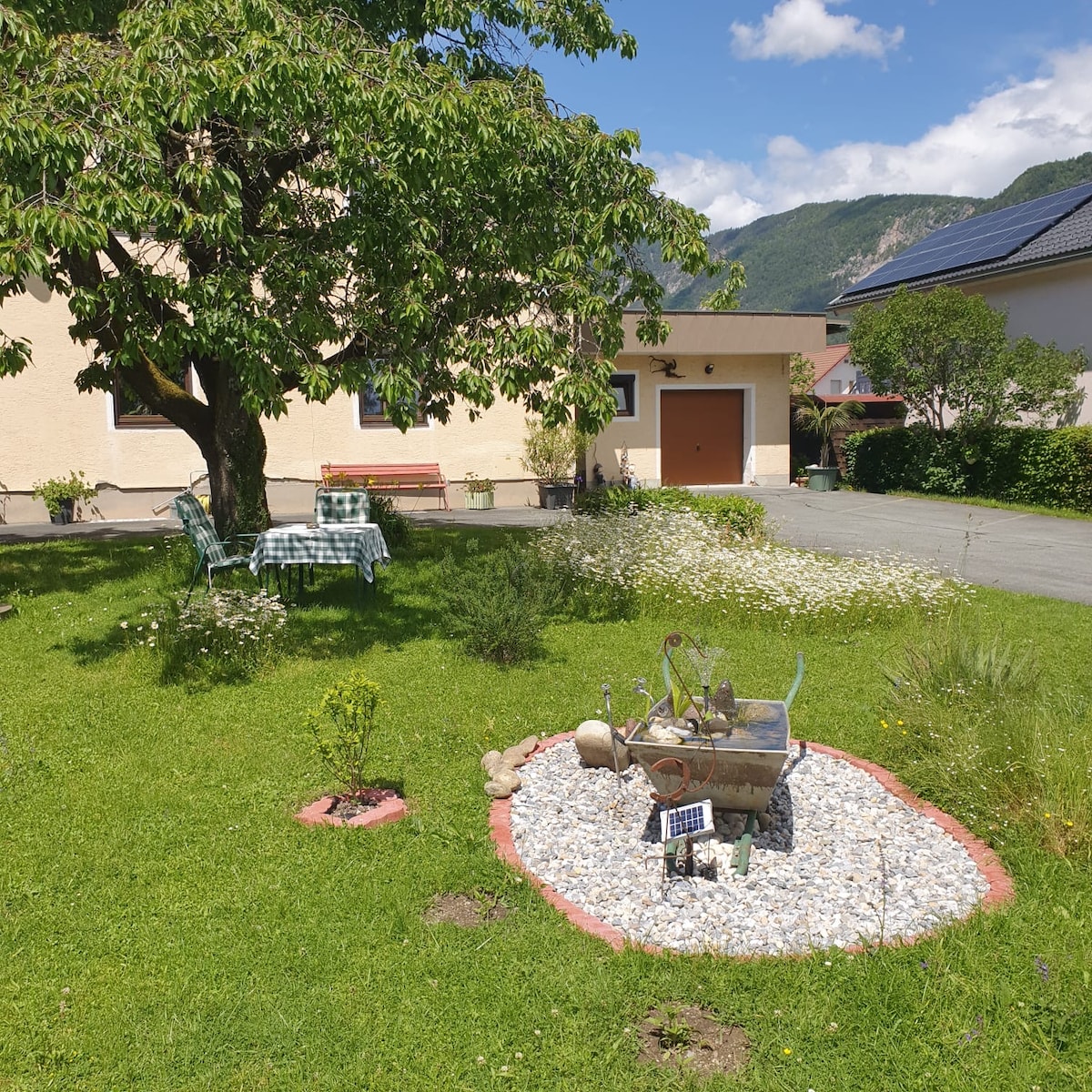 A grassy garden area features a large cherry tree providing shade, accompanied by two green chairs and a wooden bench. A small circular stone arrangement with decorative rocks is centered in the garden, offering a peaceful spot to relax amidst the surrounding greenery.