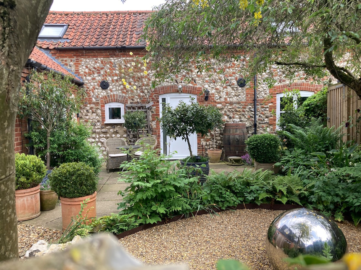 A lush courtyard garden is framed by greenery, featuring various plants and pot arrangements. Smooth pebbles create a path, leading to seating areas that include outdoor chairs. The stone building's rustic façade is visible, complemented by garden features such as a spherical sculpture.