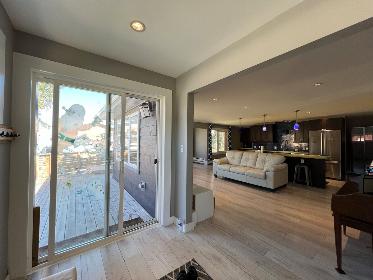 An open living space connects to a wooden deck through sliding glass doors. Natural light filters in, illuminating a light-colored sectional sofa. The kitchen area features modern appliances, with pendant lighting enhancing the overall ambiance. A neutral color palette contributes to a spacious feel.
