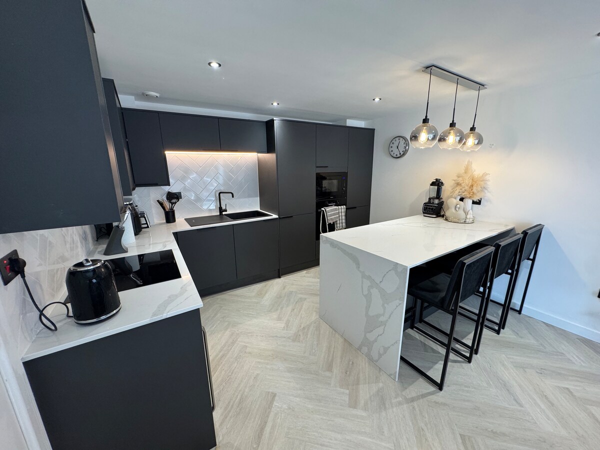 A modern kitchen is equipped with sleek black cabinetry and a spacious island featuring white countertops. Stainless steel appliances are positioned along the walls, while black stools are arranged around the island. Natural light enhances the minimalist design, completed by stylish pendant lighting above.