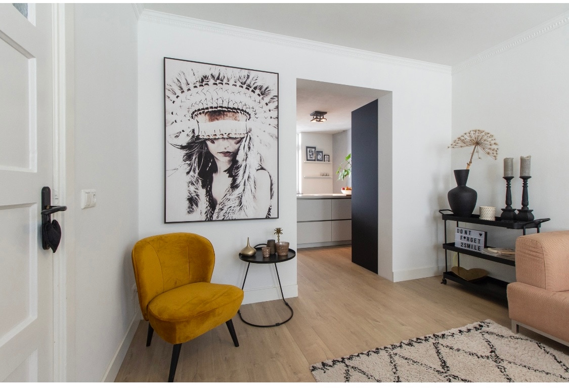 A bright and inviting entryway is characterized by a velvet armchair in warm mustard yellow. A decorative wall piece featuring a nature-inspired design adorns the space, accompanied by a small side table holding a candle. The room flows into a modern kitchen area visible through the archway.