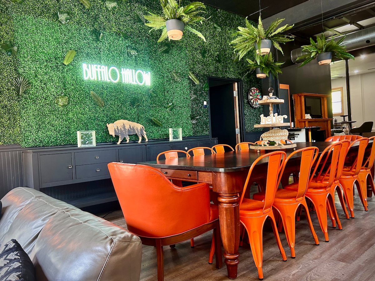 A bright dining area features a long wooden table surrounded by orange metal chairs. A wall adorned with lush greenery provides a vibrant backdrop, with decorative elements including a sign reading 'Buffalo Hallow.' Soft lighting and a dartboard enhance the inviting atmosphere.
