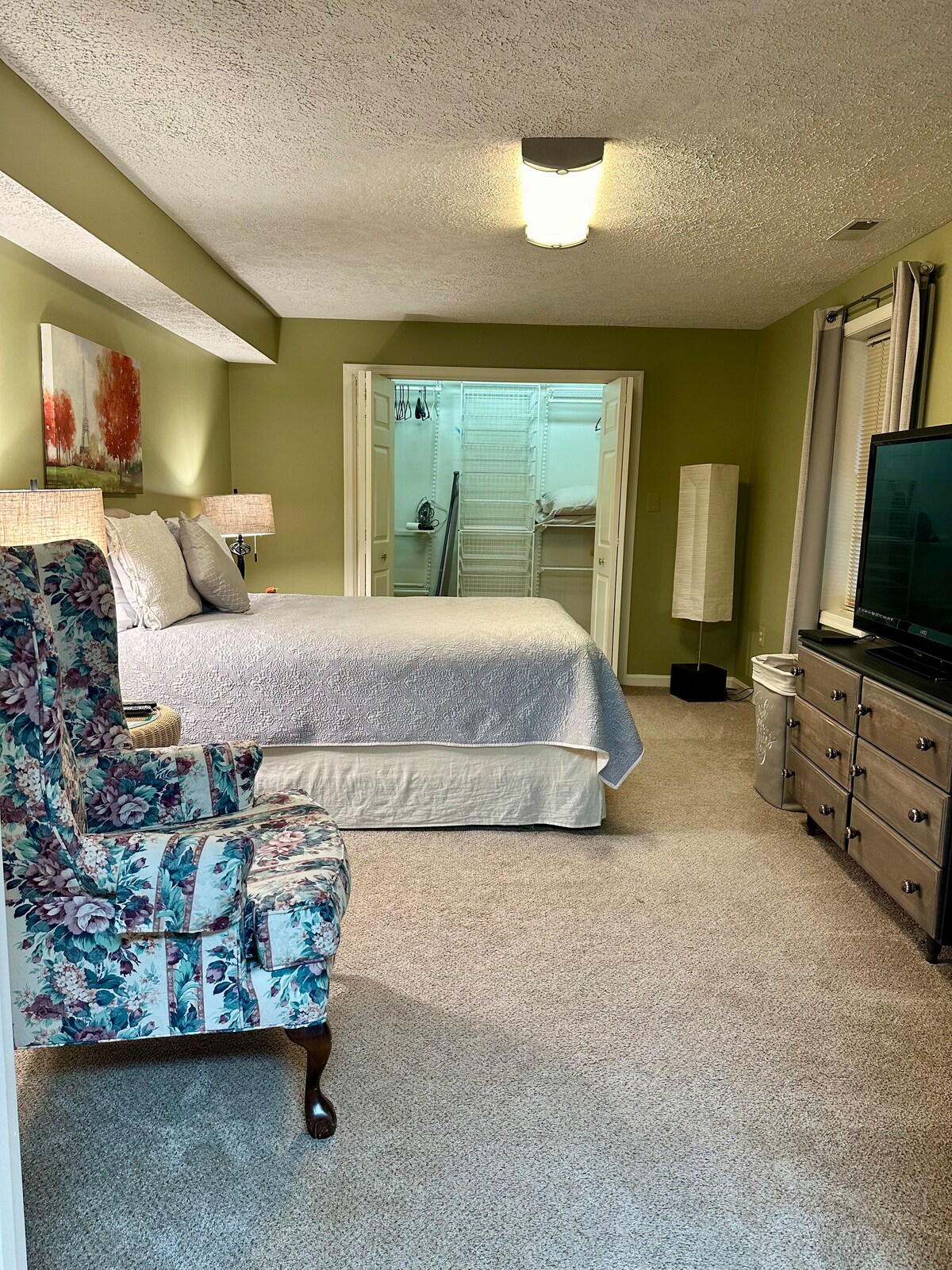 A spacious bedroom is displayed, featuring a large bed covered with a light-colored quilt. A cozy floral chair is positioned nearby, alongside a wooden dresser and television. Soft lighting enhances the calming green walls, while a closet area is visible through open double doors.