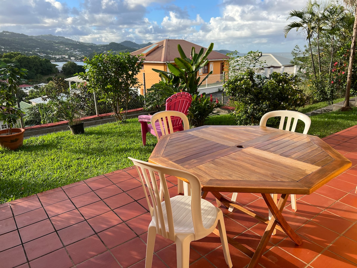 An outdoor dining area features a wooden octagonal table surrounded by three chairs, set on a red-tiled patio. The lush green garden includes tropical plants, with a view of the landscape and distant hills under a partly cloudy sky.