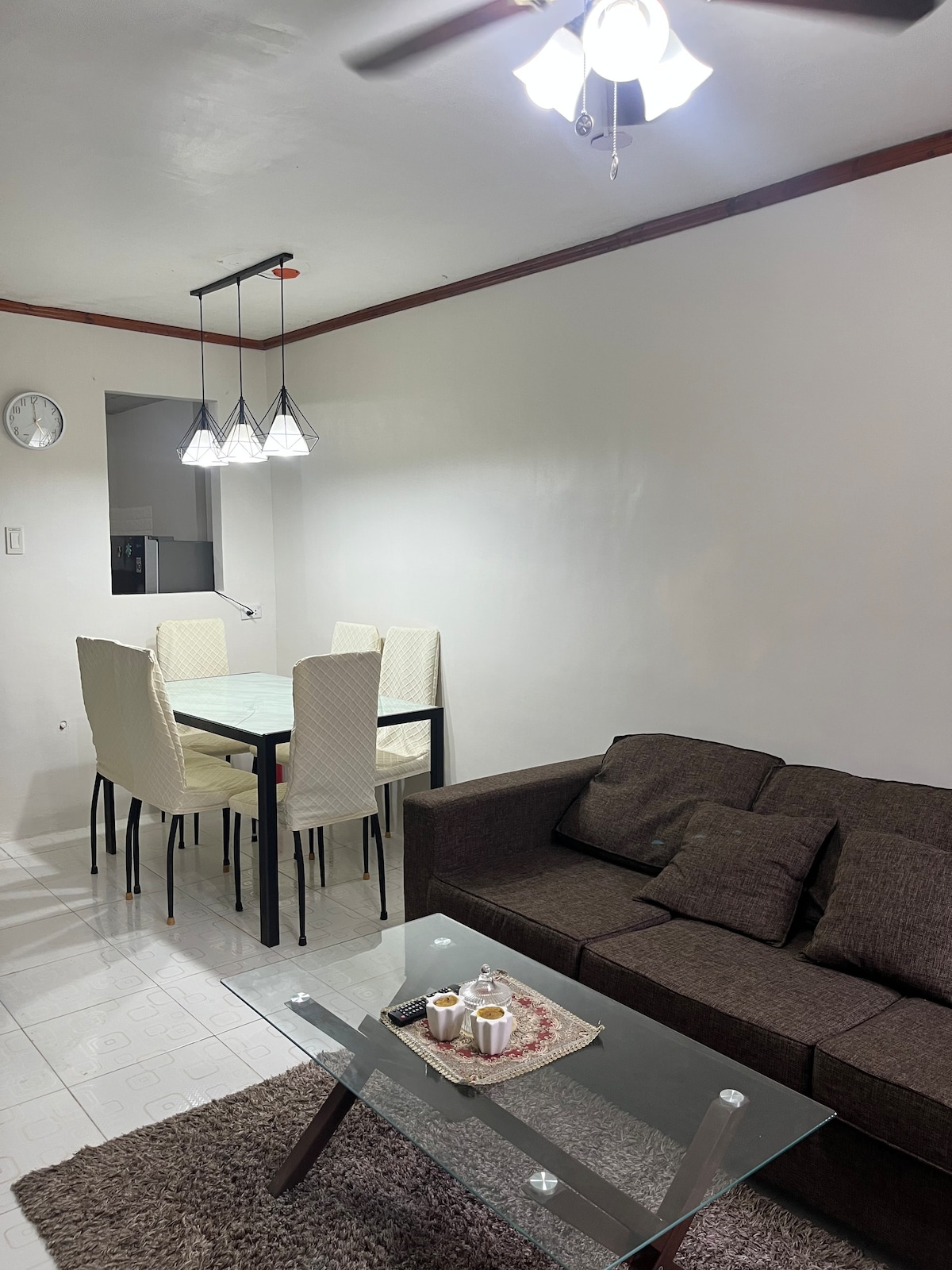 A modern living area features a comfortable brown sofa with several cushions, paired with a glass coffee table. A dining table with four chairs is positioned nearby, illuminated by pendant lighting. A wall clock is visible, and light walls create an open atmosphere.