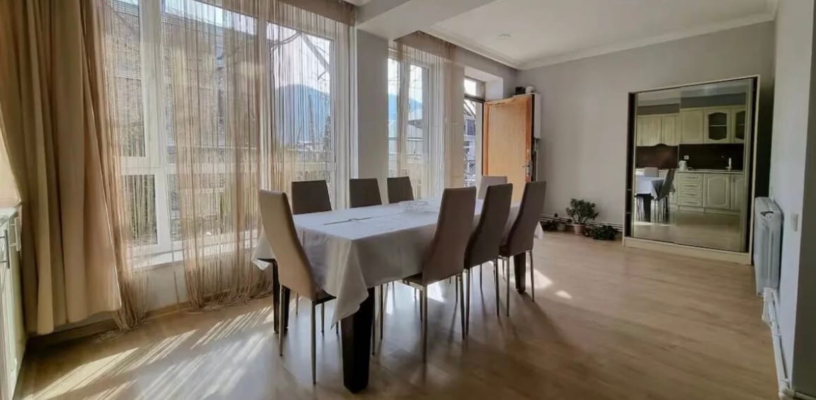 A dining area is presented with a long table surrounded by multiple chairs. Natural light filters through large windows adorned with light curtains, creating a bright atmosphere. A mirror is positioned on one wall, reflecting the room's spaciousness.