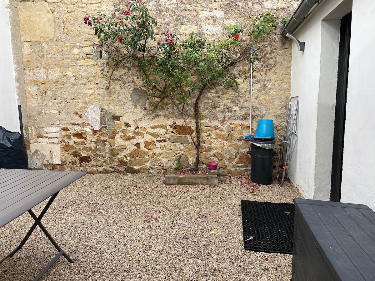 An enclosed courtyard is presented, featuring a gravel surface and a stone wall adorned with climbing greenery. A small planter is visible, along with a blue water container and a black mat near the entrance. A table is positioned to one side, providing outdoor seating.