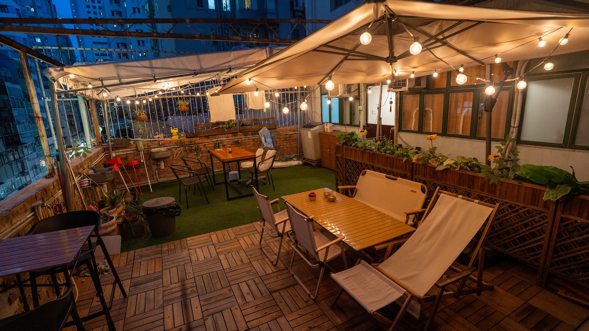 An outdoor shared space features well-equipped seating arrangements including wooden tables and lounge chairs. Soft lighting creates a cozy ambiance under a canopy, while potted plants enhance the inviting atmosphere. The area is surrounded by tall buildings, offering a blend of urban living and relaxation.