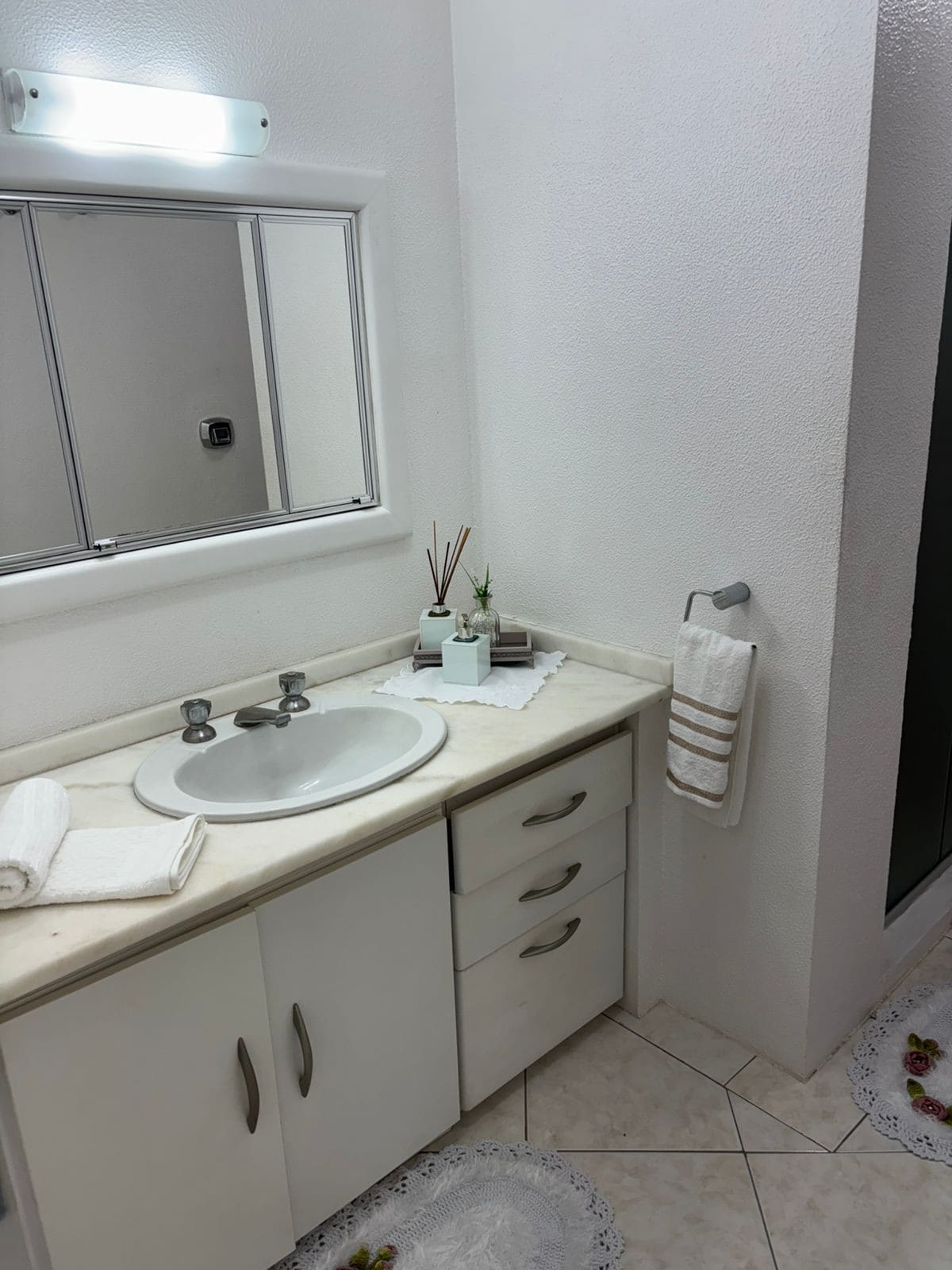 A clean bathroom is depicted with a marble countertop and a round sink featuring dual faucets. A mirror reflects the space, and a glass shower enclosure is visible. White towels and a decorative plant add a touch of simplicity to the minimalist decor.