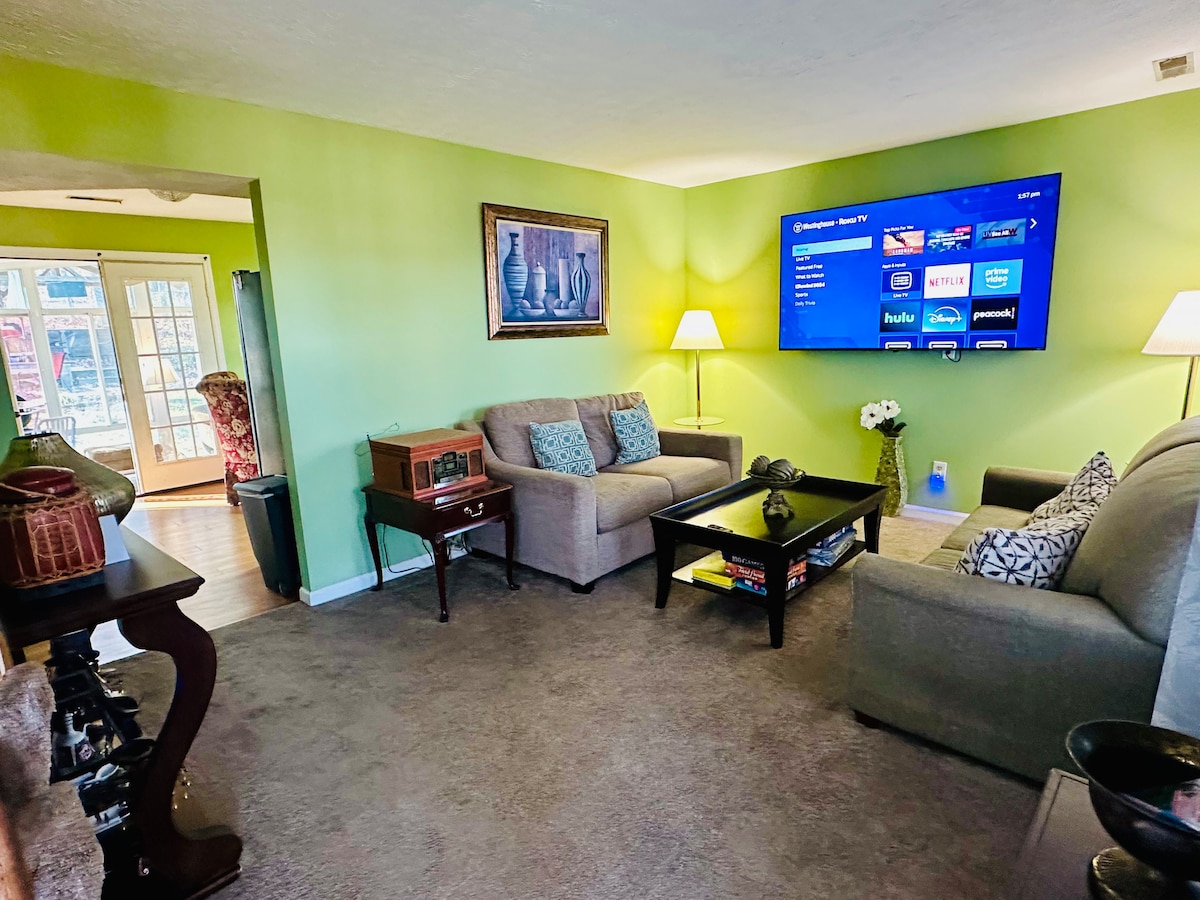 A cozy living room features soft seating arrangements with neutral-colored sofas and a contemporary coffee table. A large television mounted on a green accent wall serves as a focal point. A lamp provides additional lighting, enhancing the room's overall ambiance.