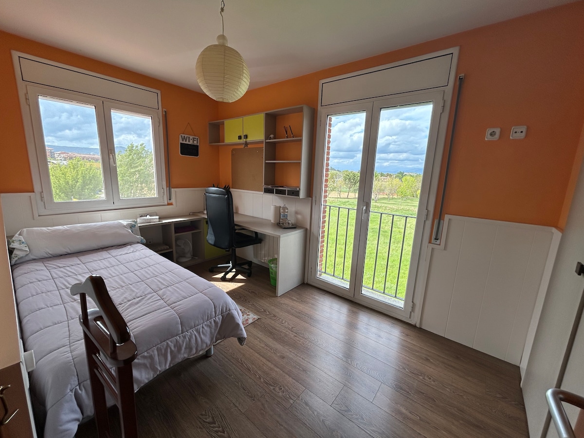 A light-filled room features an individual bed and a desk with a chair. Large windows offer views of the green landscape outside. The orange walls create a cheerful ambiance, complemented by a paper lantern hanging from the ceiling.