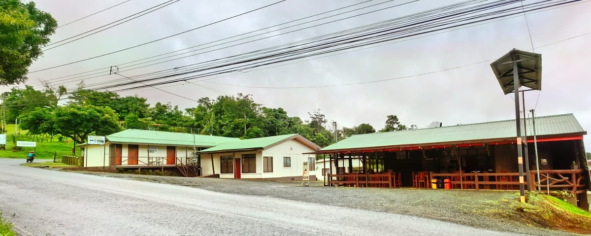 A cozy structure is surrounded by greenery, featuring a gently sloping roof and large windows. The building accommodates a welcoming entrance which is highlighted by warm wood tones. Power lines stretch above, indicating connectivity to the surrounding area.