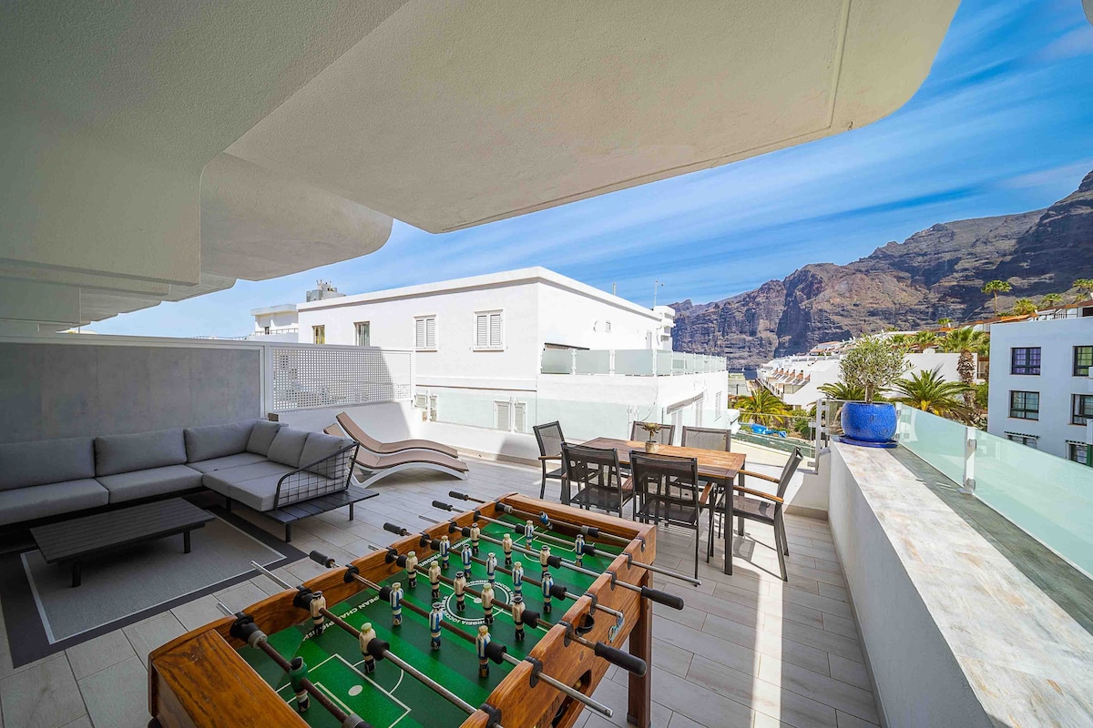 A spacious terrace features a foosball table and a dining area with a wooden table and chairs. Comfortable seating is provided by an outdoor lounge set. Scenic mountain views are visible in the background, enhancing the outdoor living space for relaxation and entertainment.