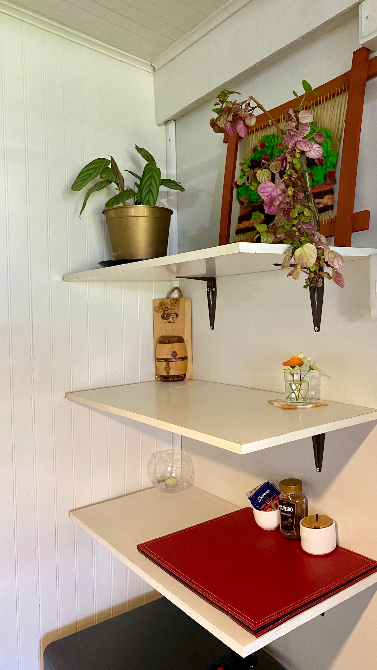 A corner shelf features a potted plant alongside a decorative wall piece. Two shelves hold a small vase with flowers, a wooden key holder, a glass globe, and various items including a notepad and a cup. The wall is painted a bright white.