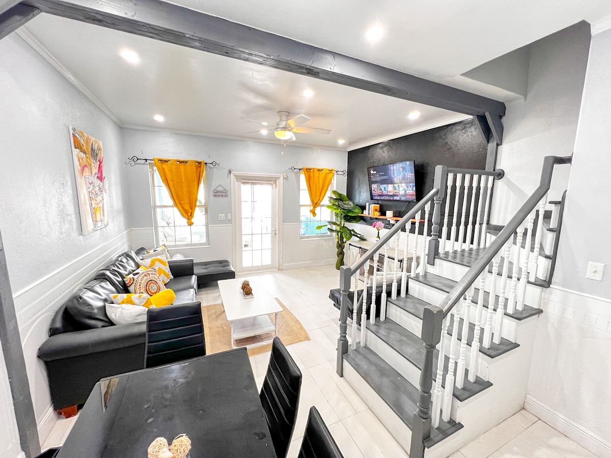 A spacious living area features a combination of modern and tropical elements, with a large sectional sofa adorned with bright yellow accents. A coffee table sits in the center, and a flat-screen TV is mounted on the wall. A staircase leads to an upper level, while natural light fills the space.