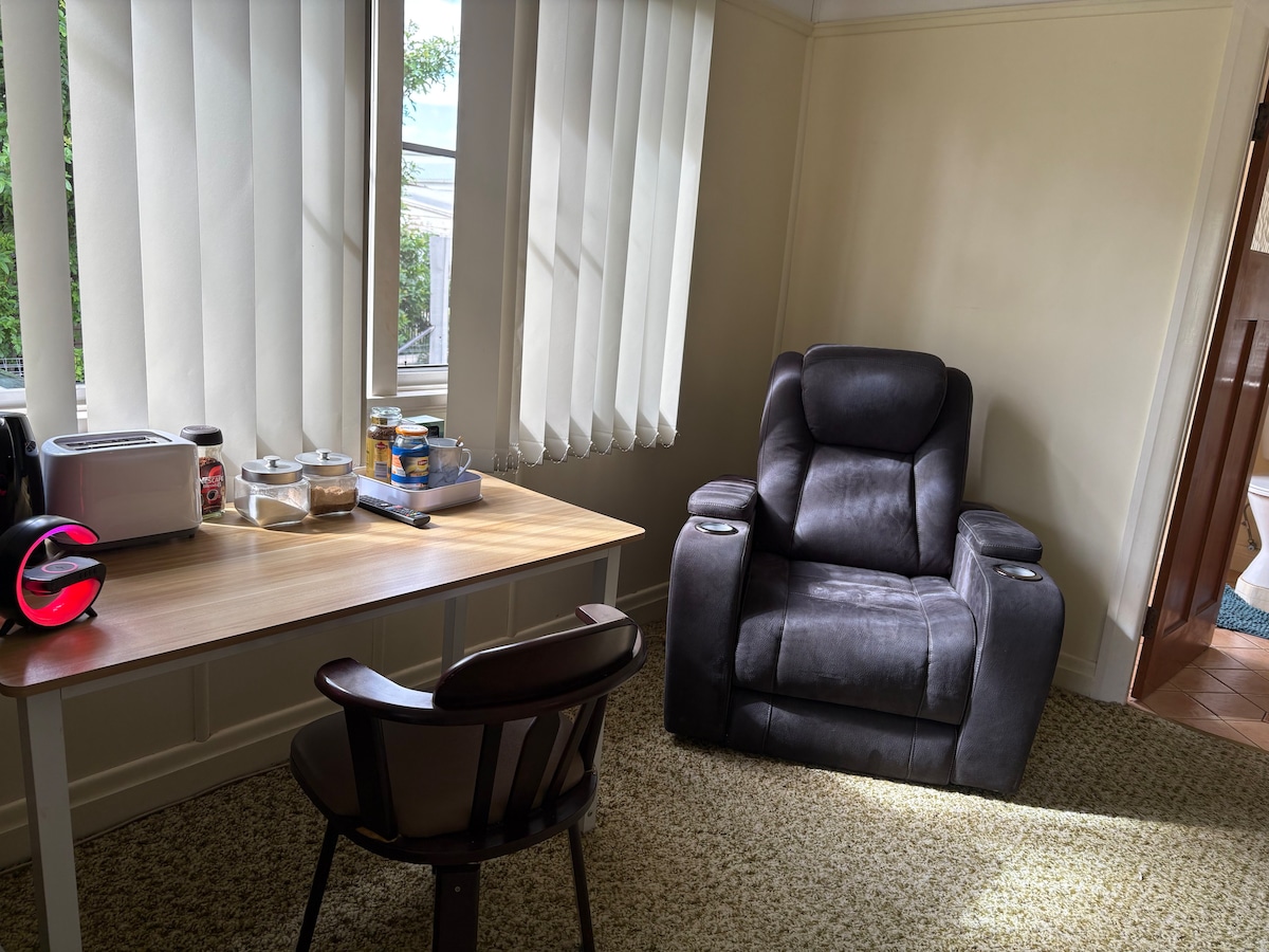 A sunlit corner features a wooden table paired with a chair, positioned near large windows adorned with vertical blinds. A plush recliner sits adjacent, creating a comfortable reading nook. Various kitchen items are organized on the table, contributing to a homey environment.