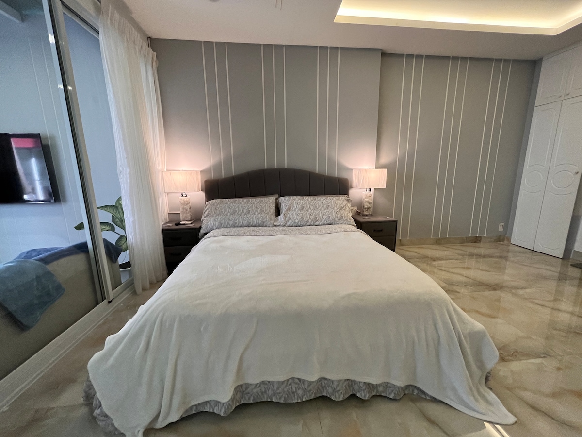 A spacious sleeping area features a large bed dressed in soft linens, flanked by two bedside lamps casting a gentle glow. A large window is partially draped with light curtains, allowing natural light to enter the room. A wall-mounted television is visible, enhancing the modern decor.