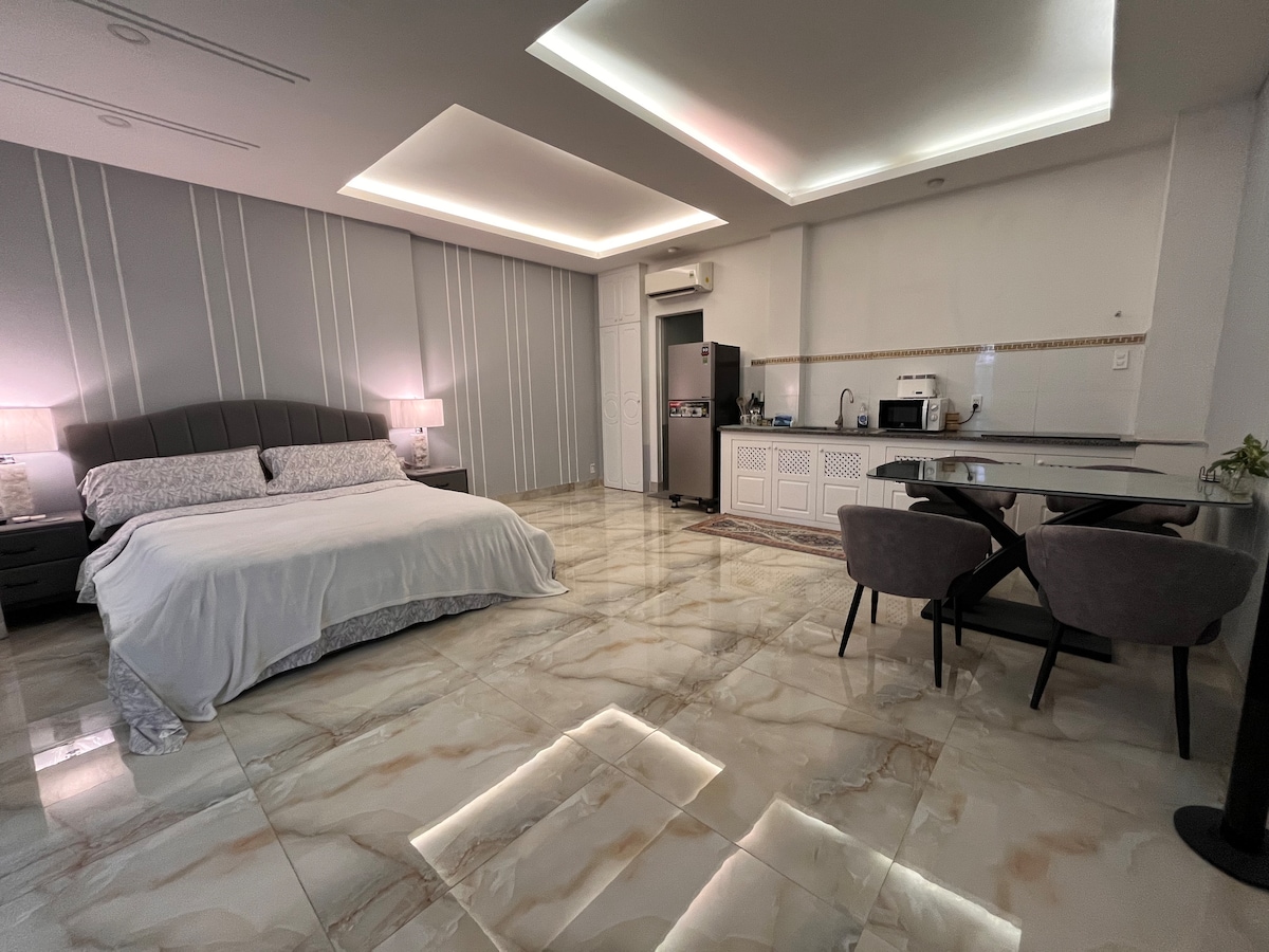 A spacious bedroom features a king-size bed with soft linens, complemented by bedside tables. An open kitchenette is visible, including a refrigerator and microwave. A dining area with sleek chairs is present, while stylish flooring reflects light, enhancing the modern design.