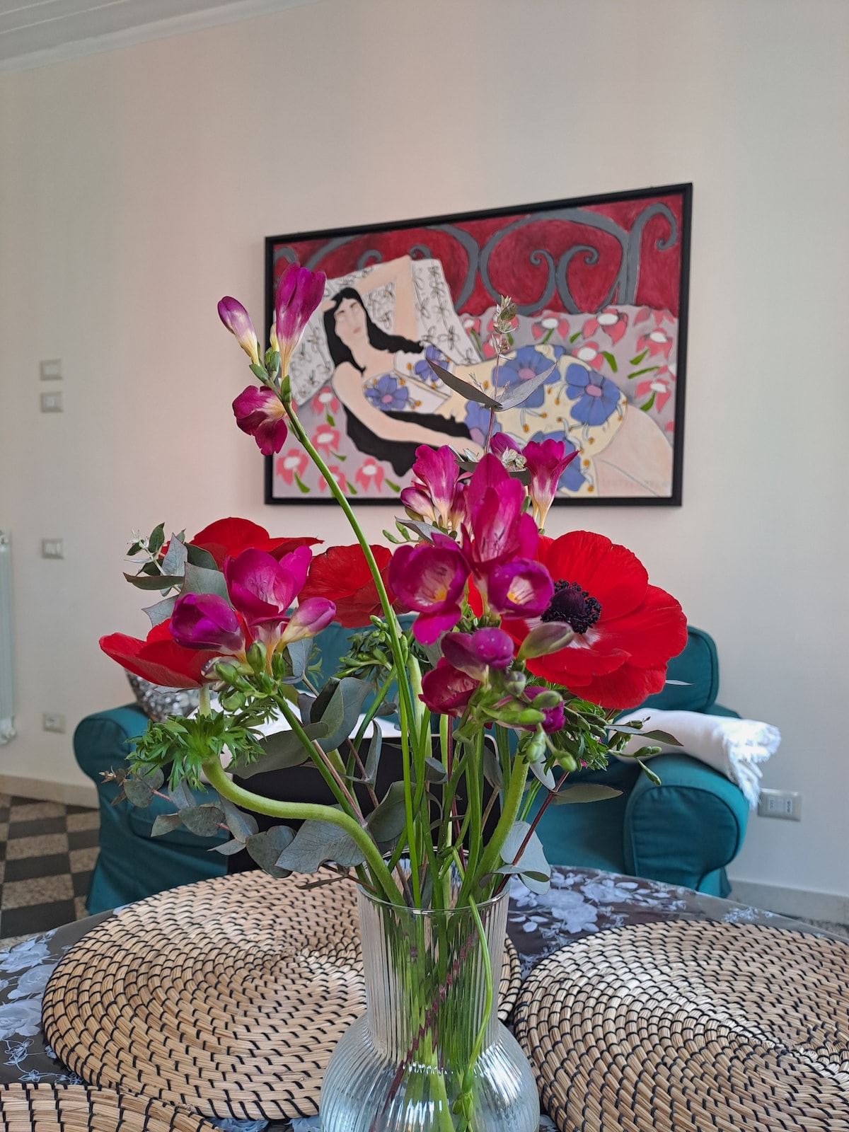 A clear glass vase holds a vibrant arrangement of pink flowers, positioned on a textured, round table. In the background, a colorful artwork adorns the wall, complementing the cozy green seating area, which invites relaxation.