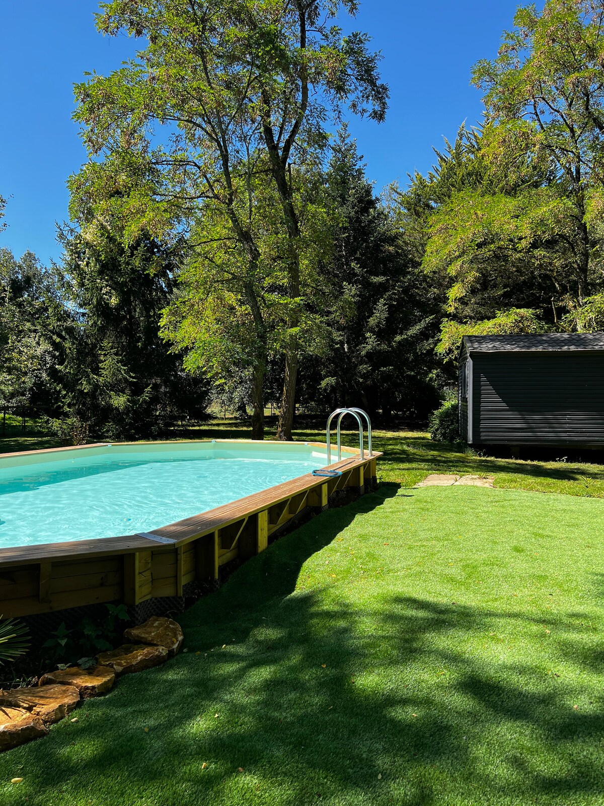 A wooden swimming pool is surrounded by lush green grass and trees, providing a natural setting. Sunlight filters through the leaves, creating dappled shadows on the ground. A small pathway leads from the pool area toward a dark-colored bungalow, enhancing the serene environment.