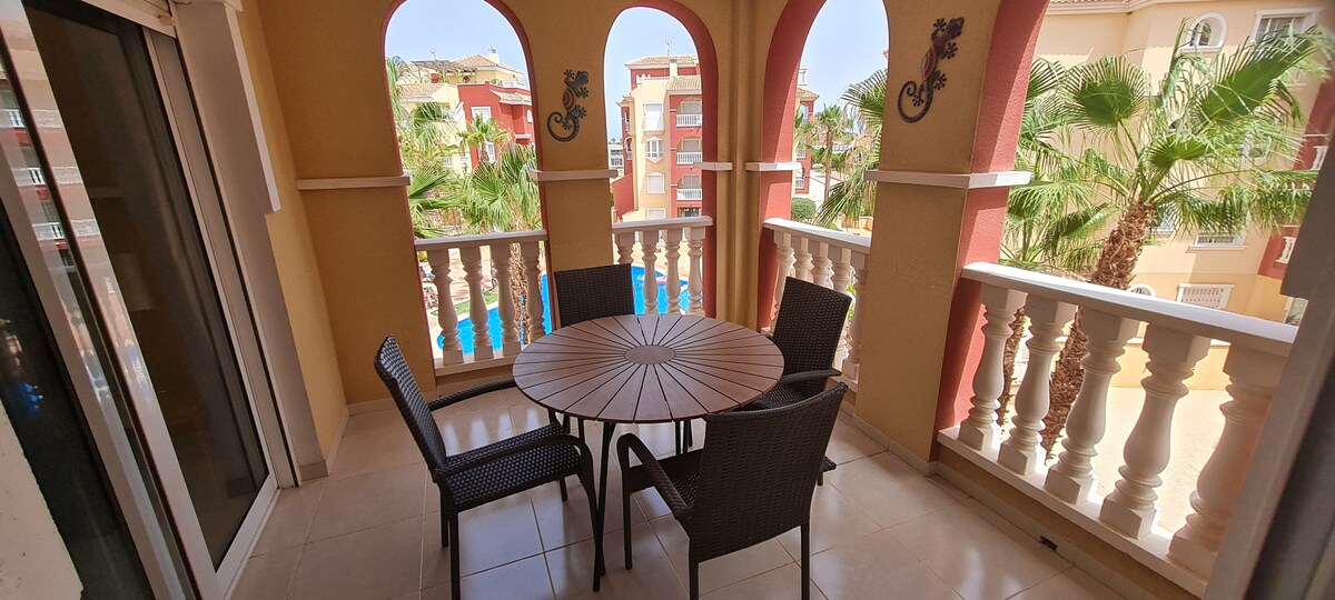A shaded balcony features a round table surrounded by four chairs. The view includes palm trees and one of the swimming pools in the distance, with arches framing the space to create a relaxed outdoor setting.