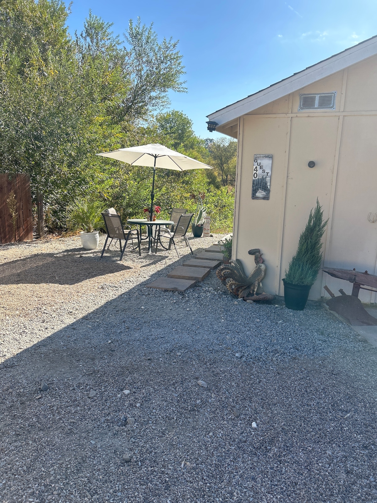 A charming outdoor seating area is visible, featuring a small table with four chairs under a large umbrella. Decorative plants and a gravel pathway lead to the entrance of the studio, while trees and greenery provide a natural backdrop.