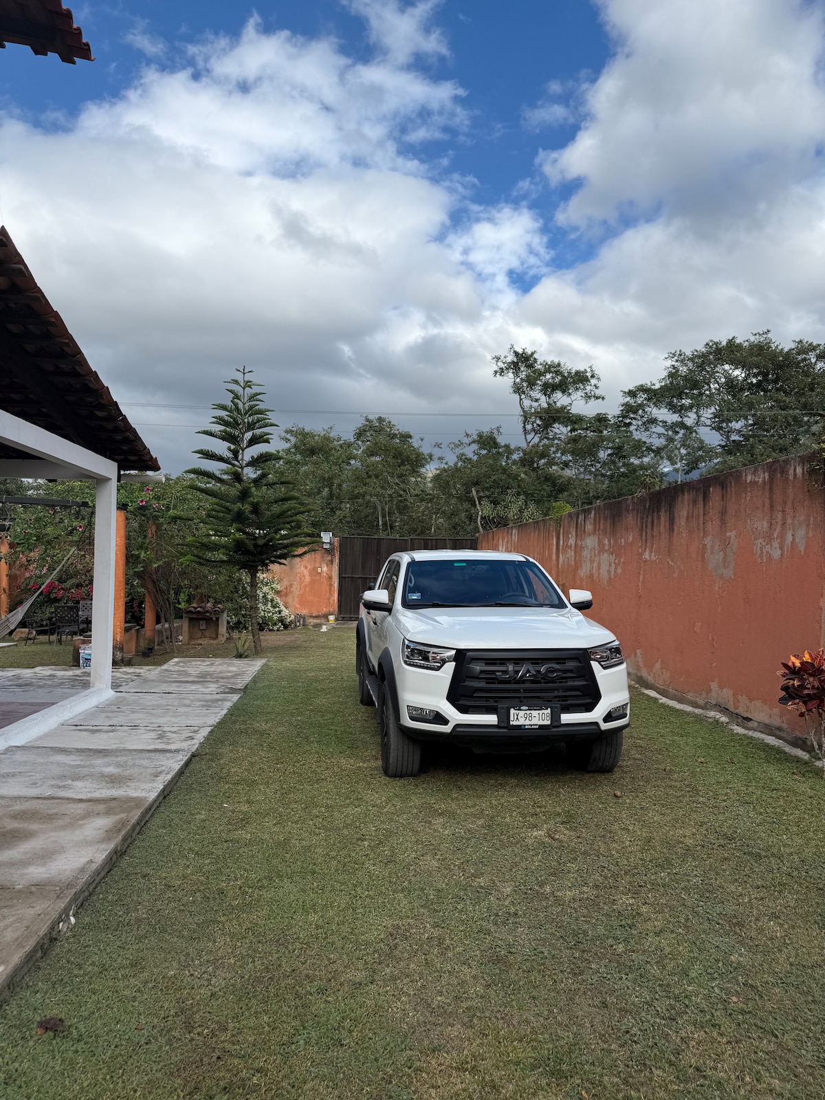 A well-maintained grassy area is visible, featuring a white pickup truck parked on the lawn. In the background, trees and a garden area are partially seen, with an orange wall surrounding the property. The sky appears partially cloudy, contributing to the outdoor ambiance.