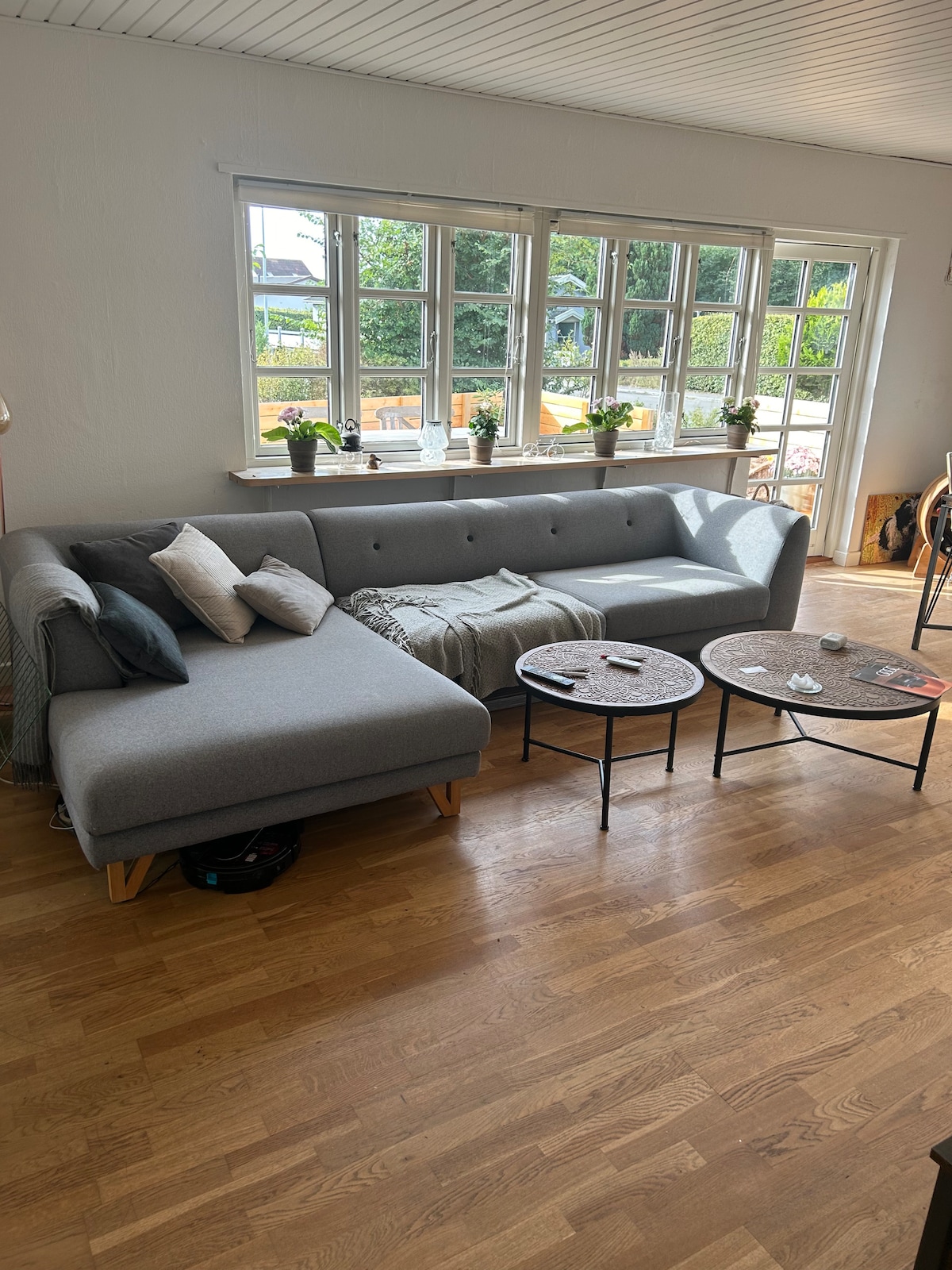 A modern sectional sofa in a light grey fabric is positioned in a bright living area. Two small, round coffee tables are placed nearby, with decorative items visible on the surfaces. Large windows allow natural light to fill the room, with greenery seen outside.