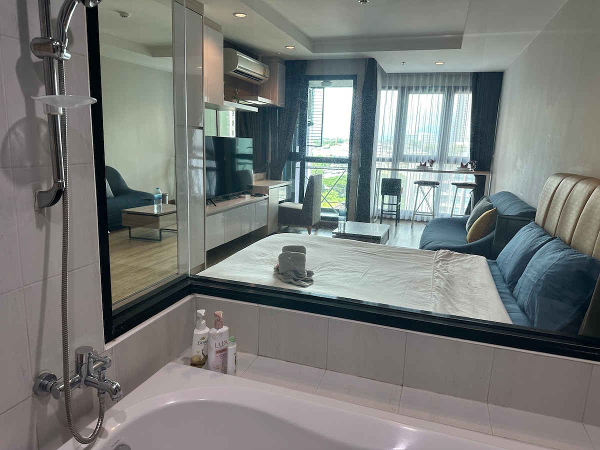 A modern bathroom is visible through a large glass window, showcasing a bathtub and toiletries on the edge. In the background, a spacious living area features a sofa and a 55-inch TV, with natural light streaming in from the large windows.