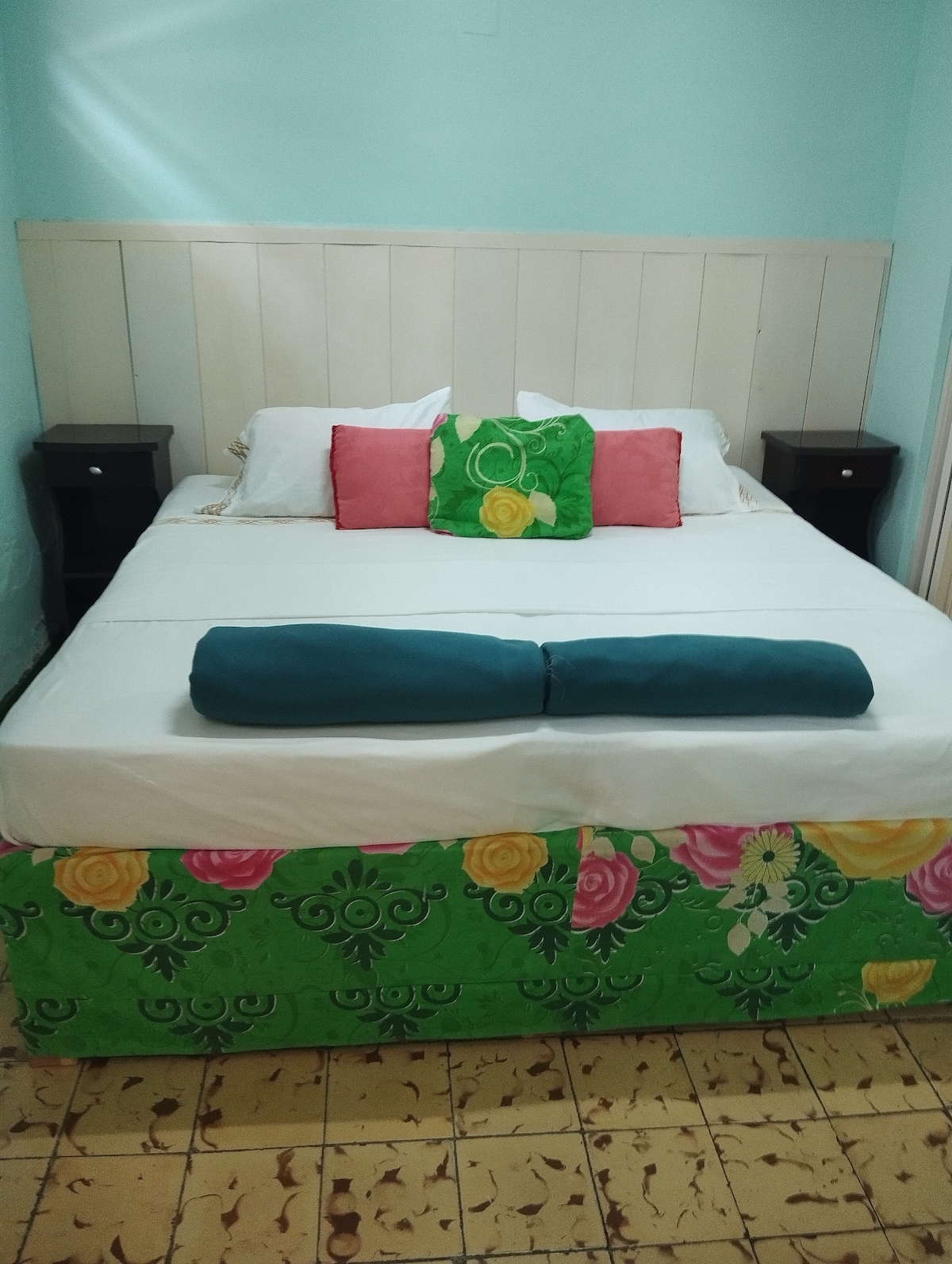A cozy sleeping area features a spacious bed with a floral-patterned bedspread. Brightly colored pillows in pink and green add a cheerful touch. Two dark nightstands are positioned on either side, complementing the light blue walls and creating a welcoming atmosphere.
