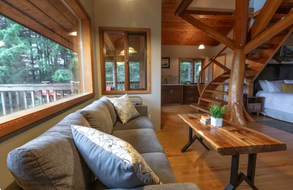 A spacious living area is highlighted by a comfortable gray couch adorned with decorative pillows. A wooden coffee table sits in front, and large windows allow natural light to illuminate the space. A spiral staircase leads to the upper level, visible in the background.