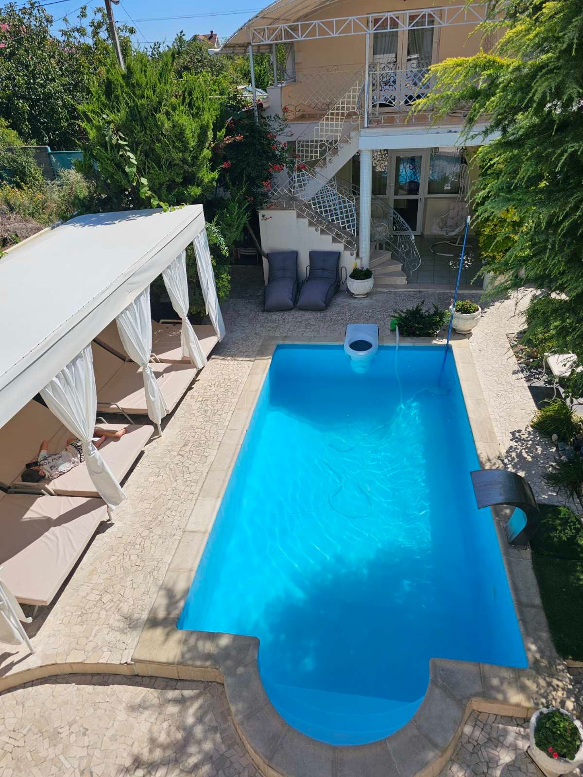 A serene outdoor area features a rectangular swimming pool with clear blue water, surrounded by greenery. Comfortable loungers are positioned under shaded cabanas, while a staircase leads to the upper level of the property. The inviting space is designed for relaxation and recreation.